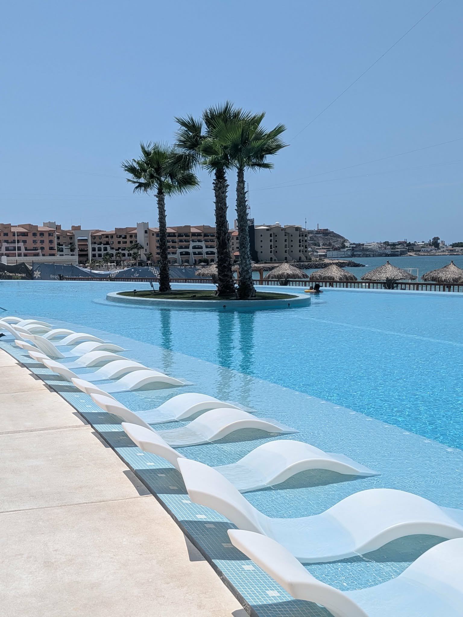 Large blue resort infinity pool with in-water loungers, palms and ocean